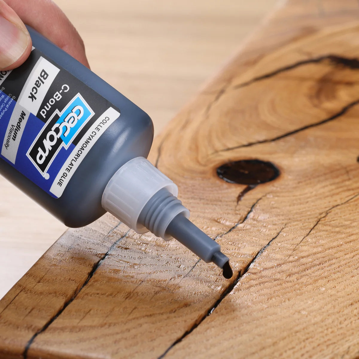 A close-up of a hand applying black wood filler or epoxy resin from a bottle labeled 'C-Bond Black' onto a wood grain surface.