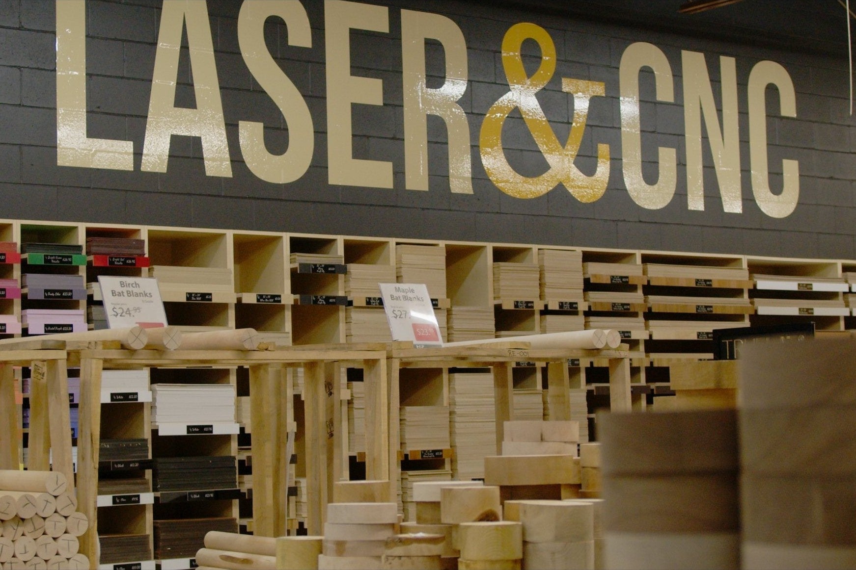 The Laser and CNC bay at The Wood Vault: the gold LASER & CNC wall lettering above open birch shelving stacked with craft plywood, acrylic sheets and blanks